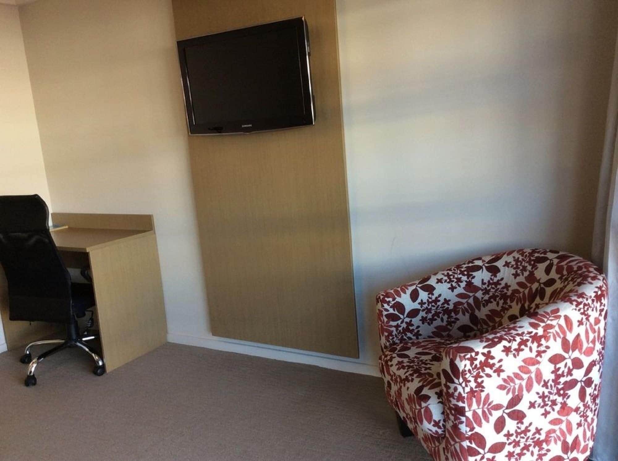 Habitación de hotel con televisor de pantalla plana montado en la pared, un escritorio con una silla y un sofá estampado con motivos florales.