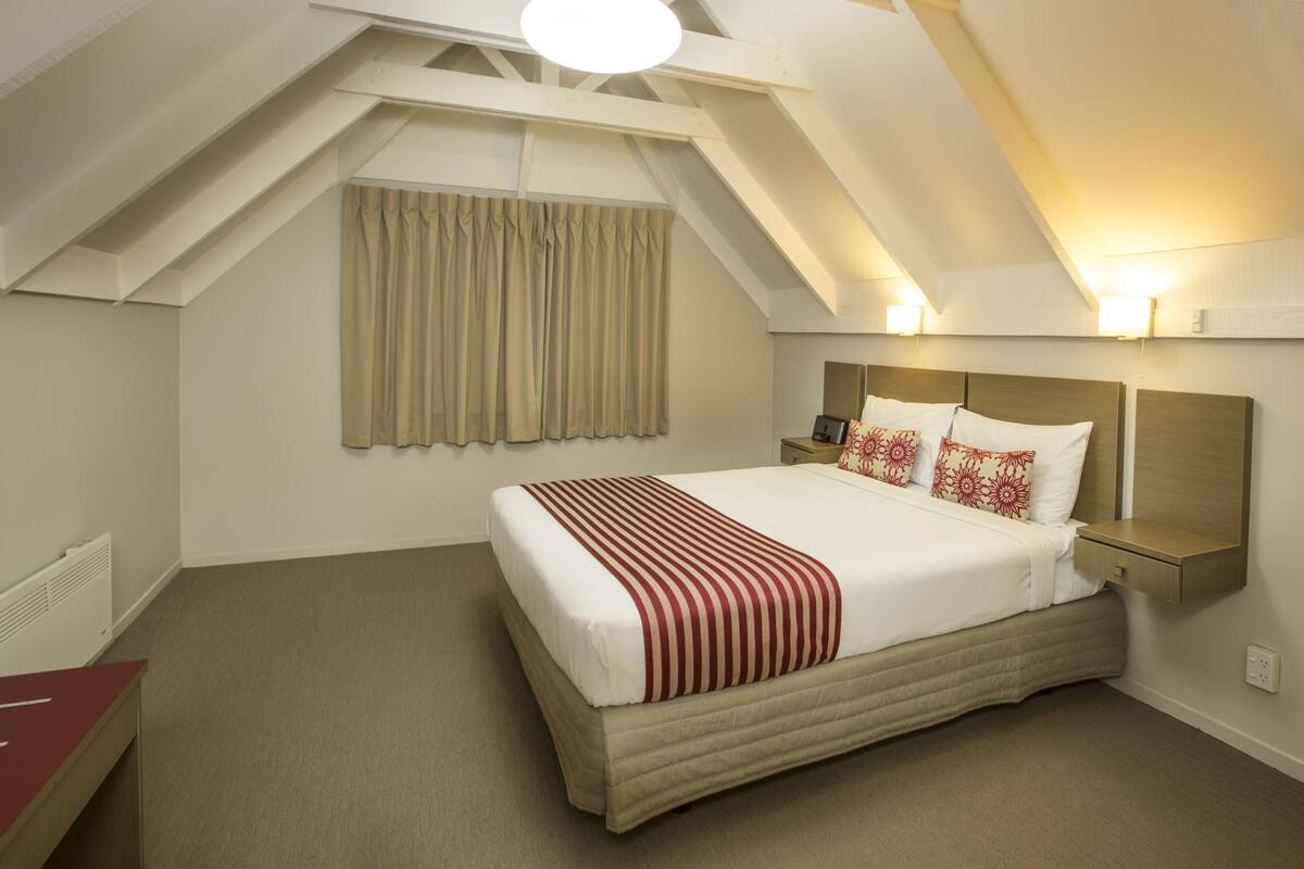 Quarto de hotel com uma cama grande, uma mesa de cabeceira, uma luminária na parede e uma janela com cortinas.