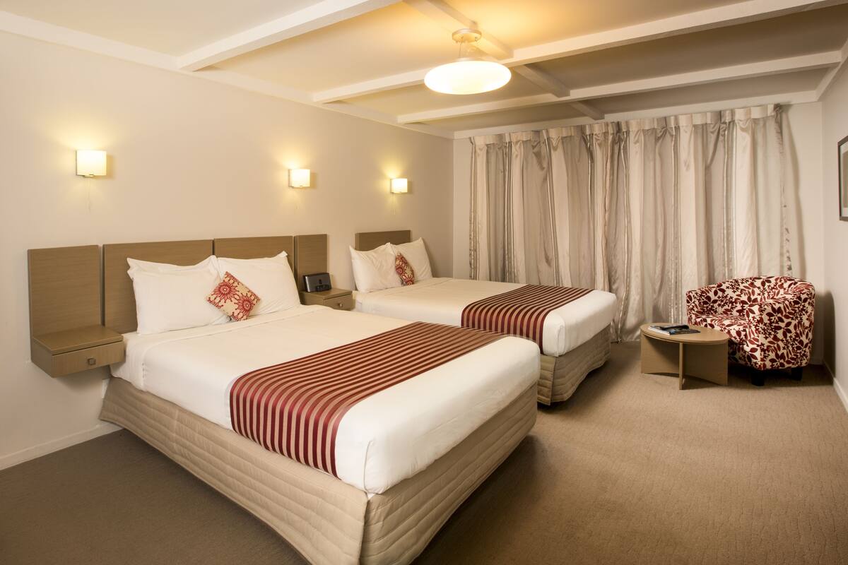 Quarto de hotel com duas camas, uma mesinha, uma cadeira e uma janela com cortinas.