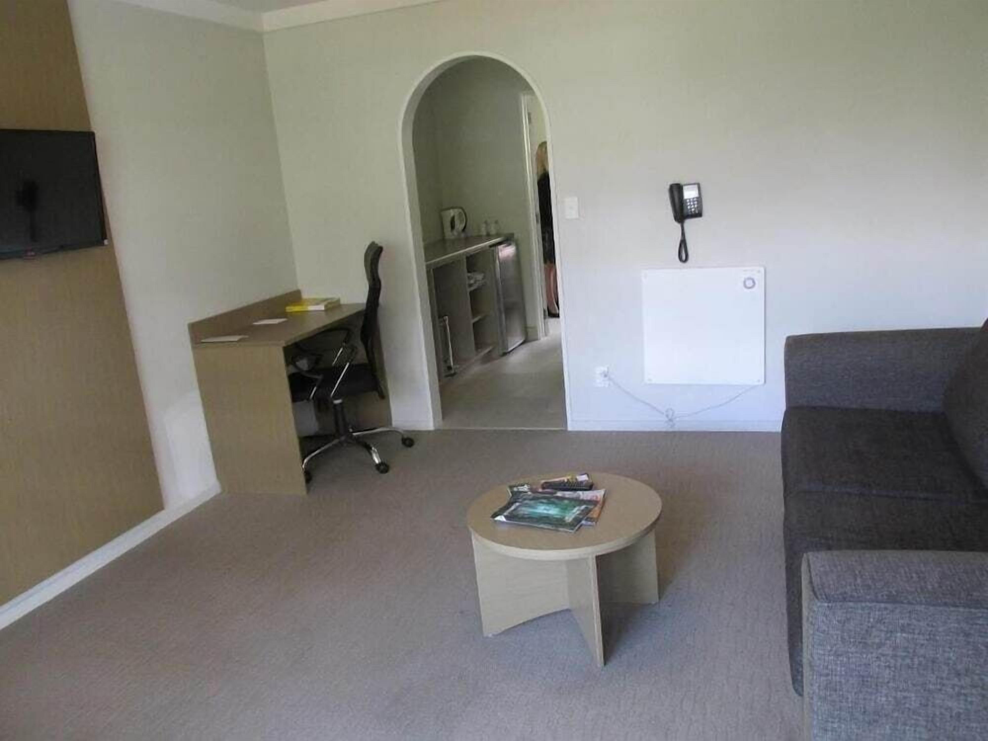 A living room with a grey sofa, a small round table with magazines, a desk with a chair, and a phone on the wall.