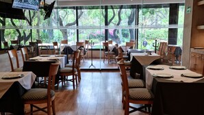 Un restaurante con suelo de madera, mesas con sillas y grandes ventanales que dan a árboles y vegetación.