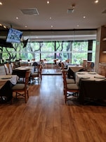 Un restaurante con suelo de madera, mesas con sillas y grandes ventanales que dan a árboles y vegetación.