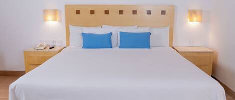 Una cama bien tendida con ropa blanca y dos almohadas azules, flanqueada por mesitas de noche de madera con lámparas y un teléfono.