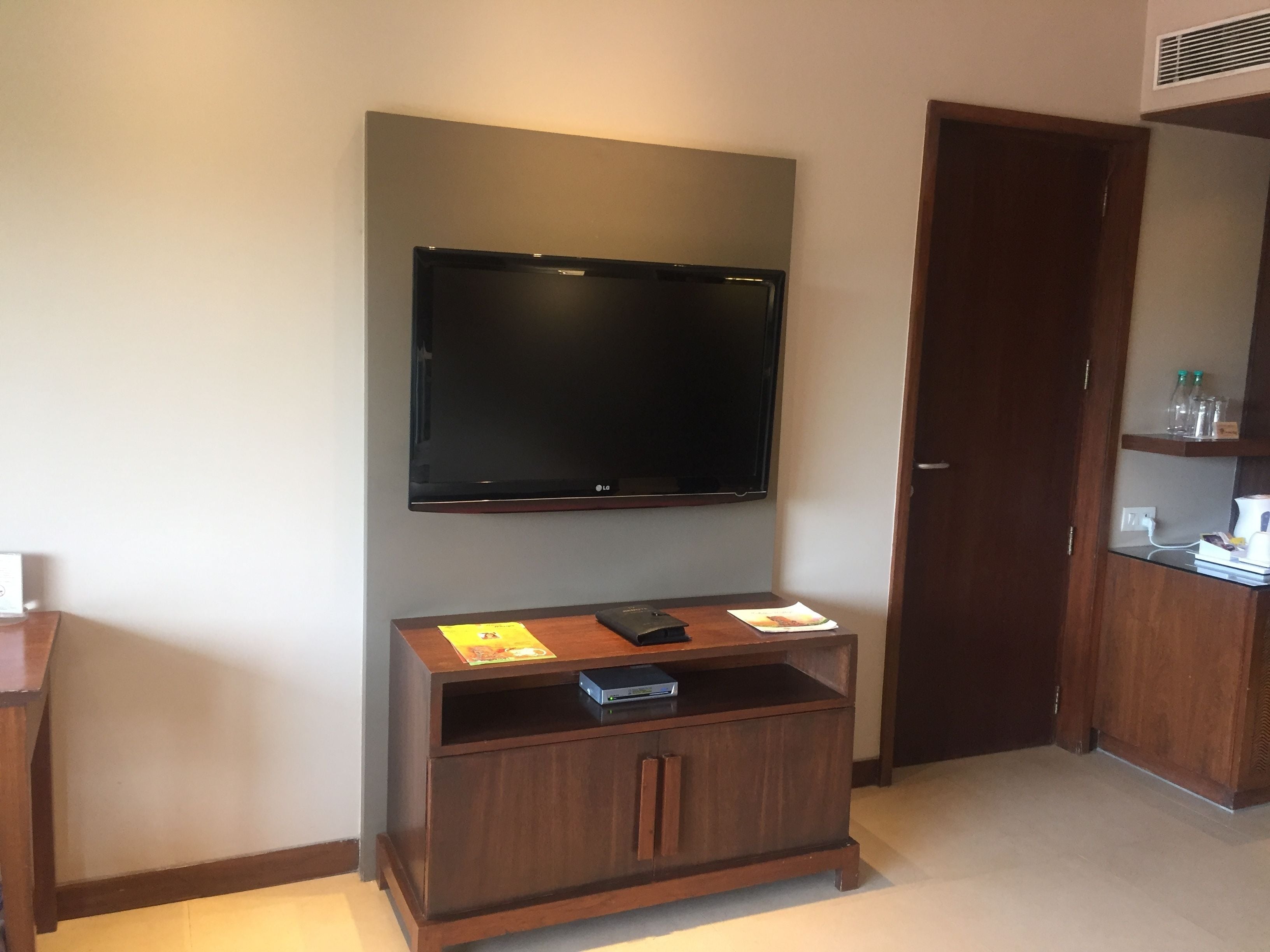 A hotel room with a flat-screen TV mounted on the wall, a wooden entertainment unit with a DVD player, and a door with a wooden frame.