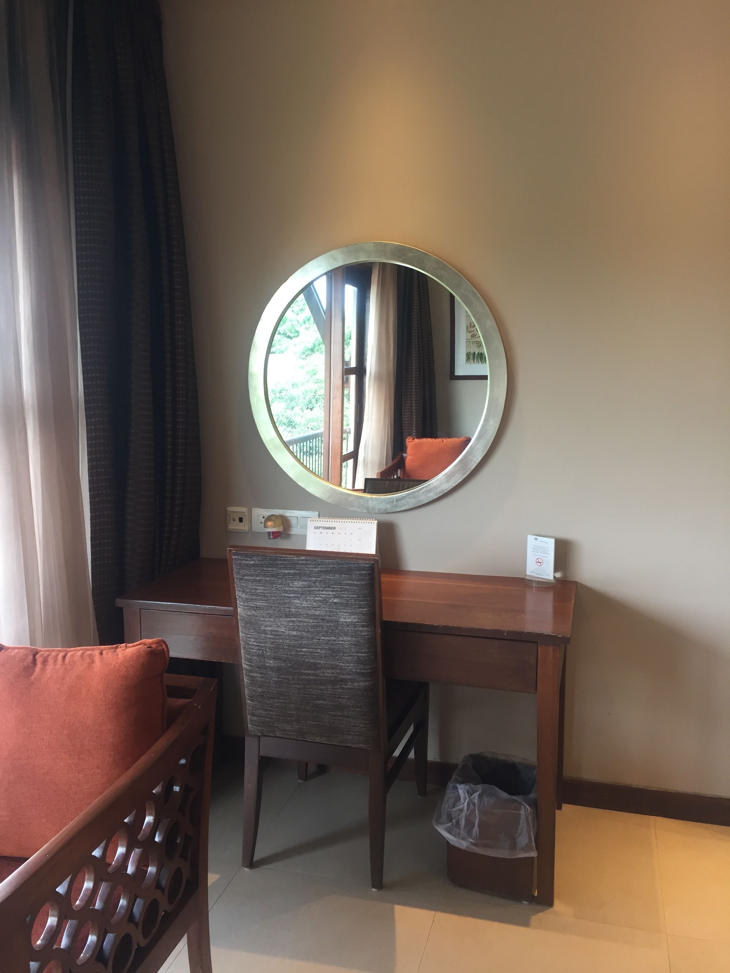 A hotel room with a round mirror, a wooden desk, a chair with a cushion, a calendar, and a trash bin.