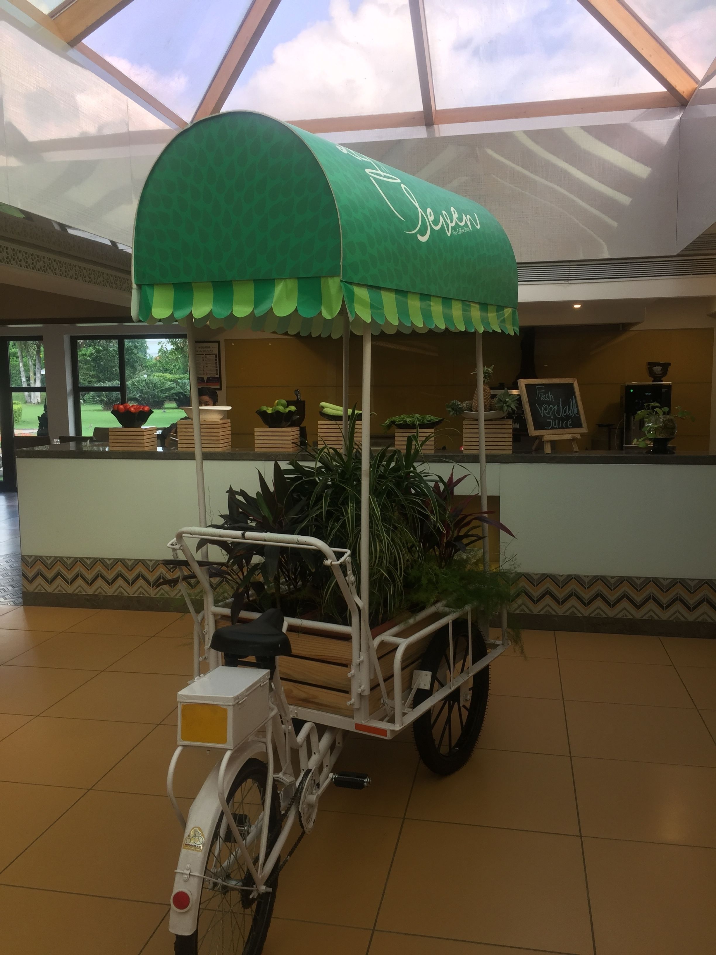 A stationary food cart with a green canopy, a white bicycle, and a sign that reads "Fresh Vegetable Juice".