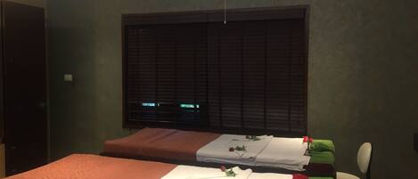 A room with two beds, each with a pillow and a rose. There is a window with blinds, a chair, and a small table.