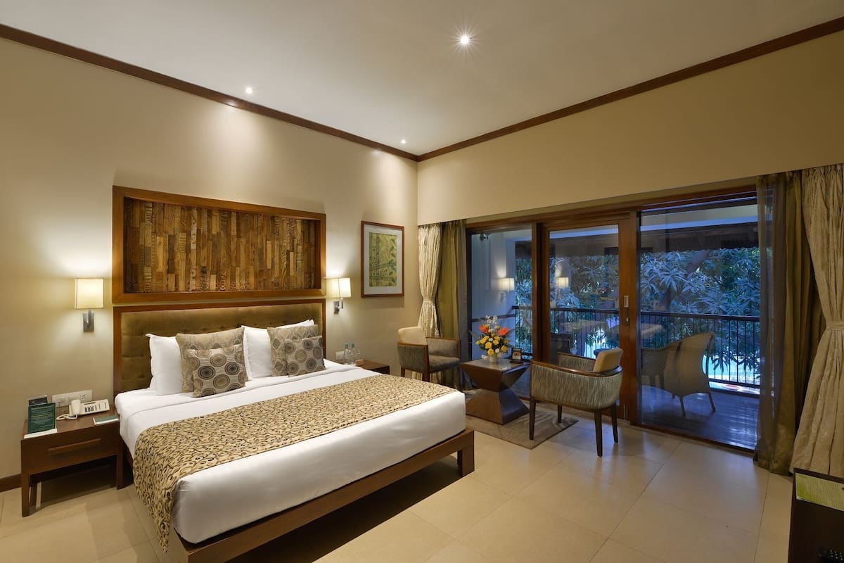 Une chambre spacieuse avec un grand lit, une tête de lit en bois, une table de chevet, une commode, une chaise, une petite table avec un vase et un balcon offrant une vue sur les arbres.