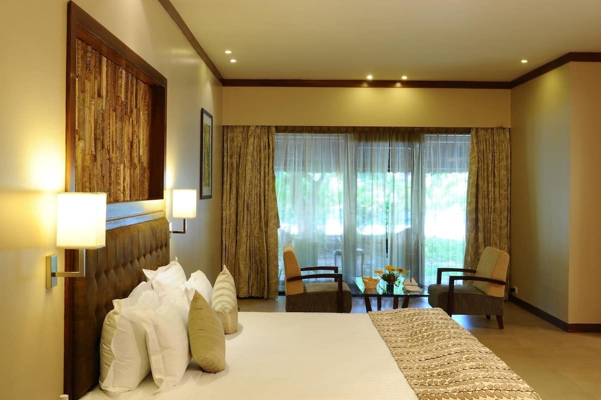 Une chambre d’hôtel avec un grand lit, deux fauteuils, une petite table et une vue sur la verdure par les fenêtres.
