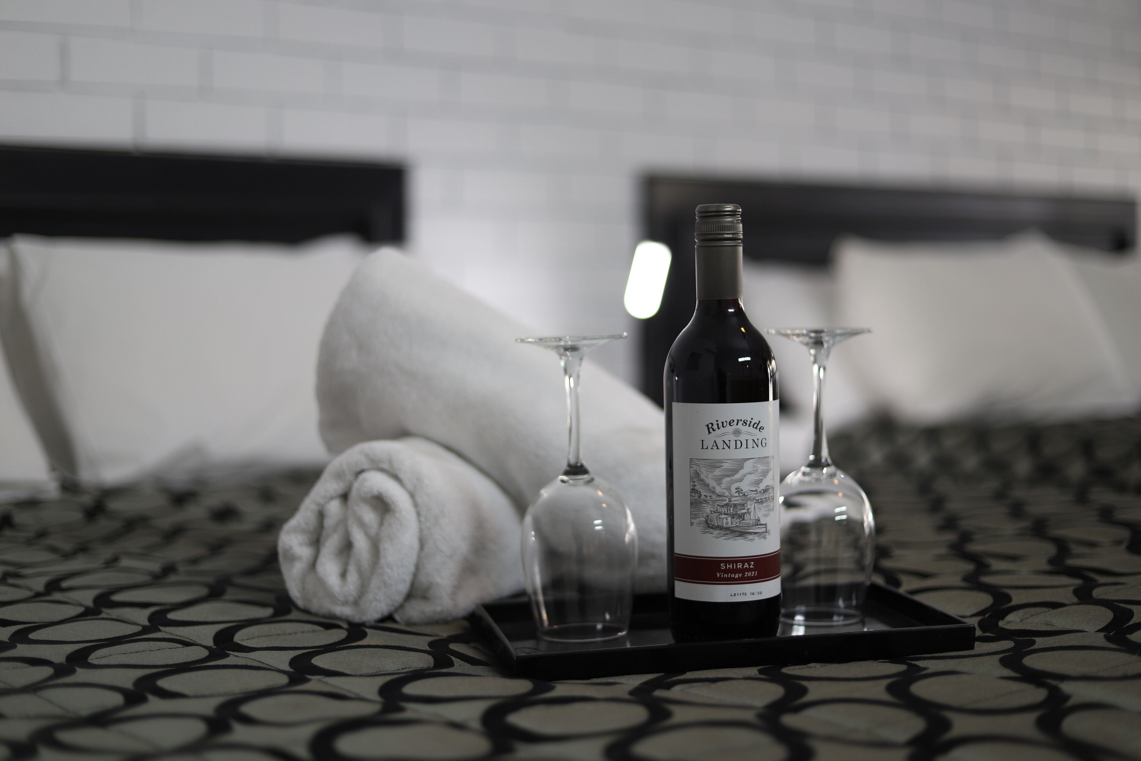 A bottle of wine and two glasses on a tray on a bed with a patterned bedspread.