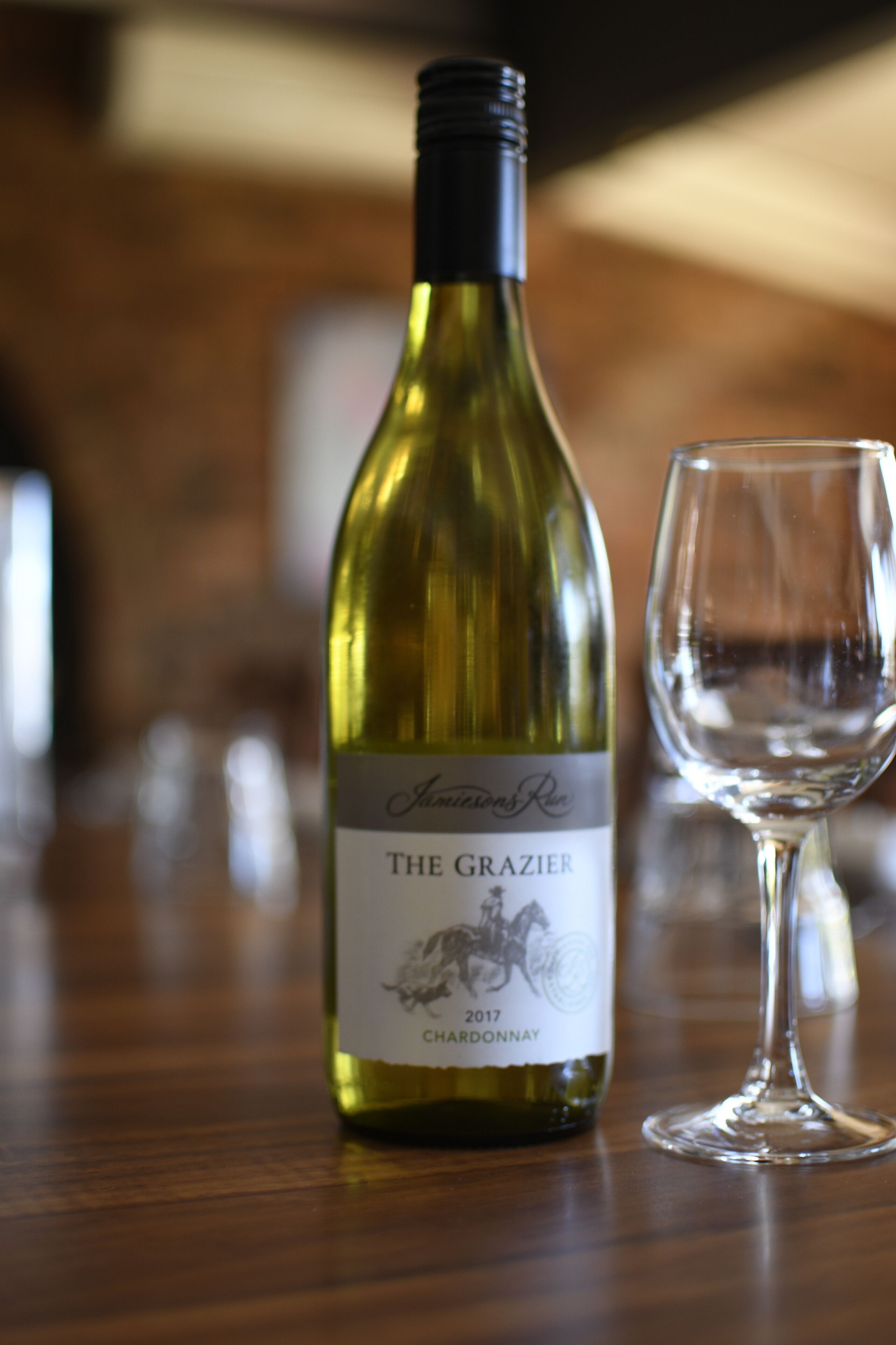 A bottle of The Grazier Chardonnay wine and an empty wine glass on a wooden surface.