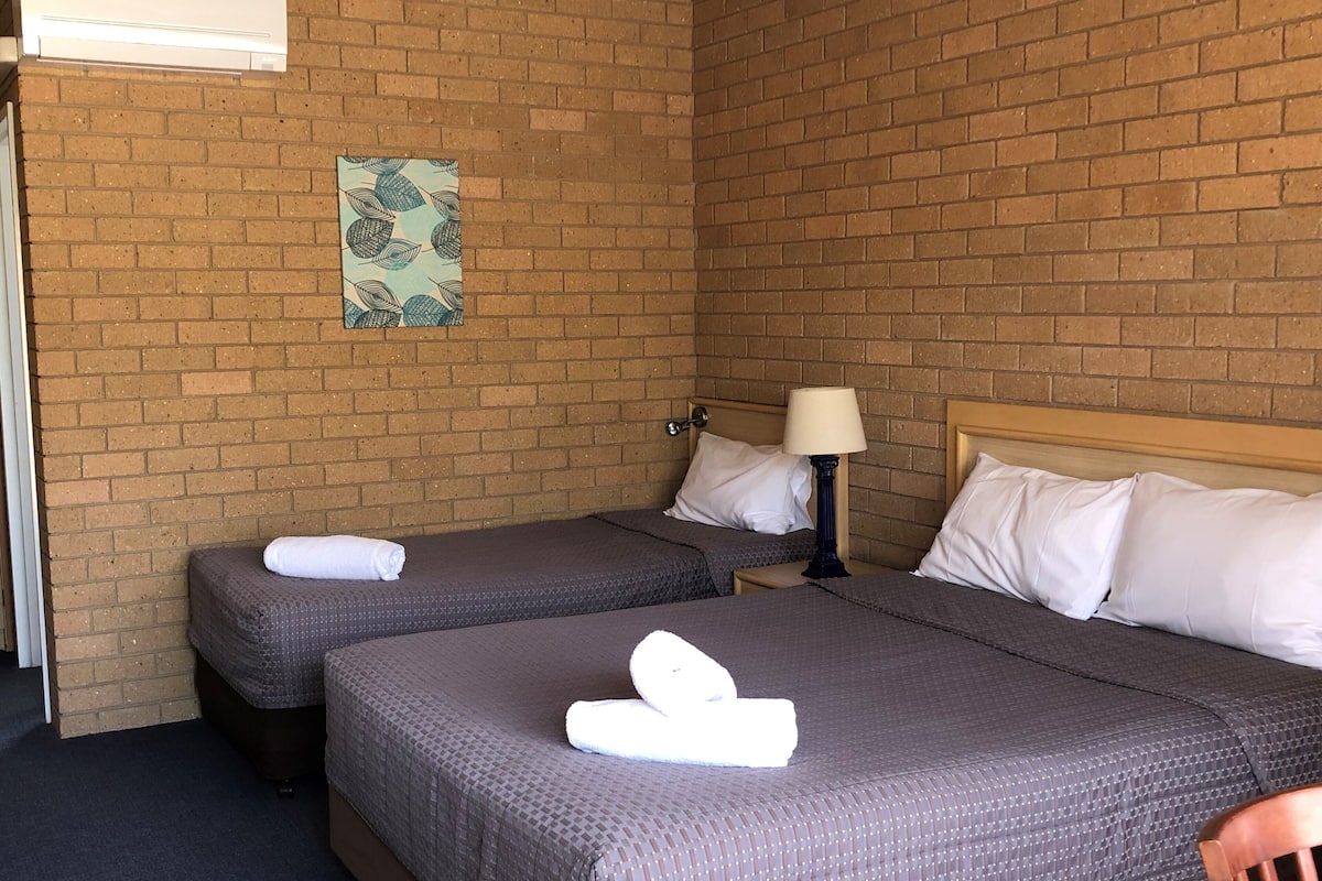 A hotel room with two beds, a brick wall, a small table with a lamp, and a decorative wall hanging.