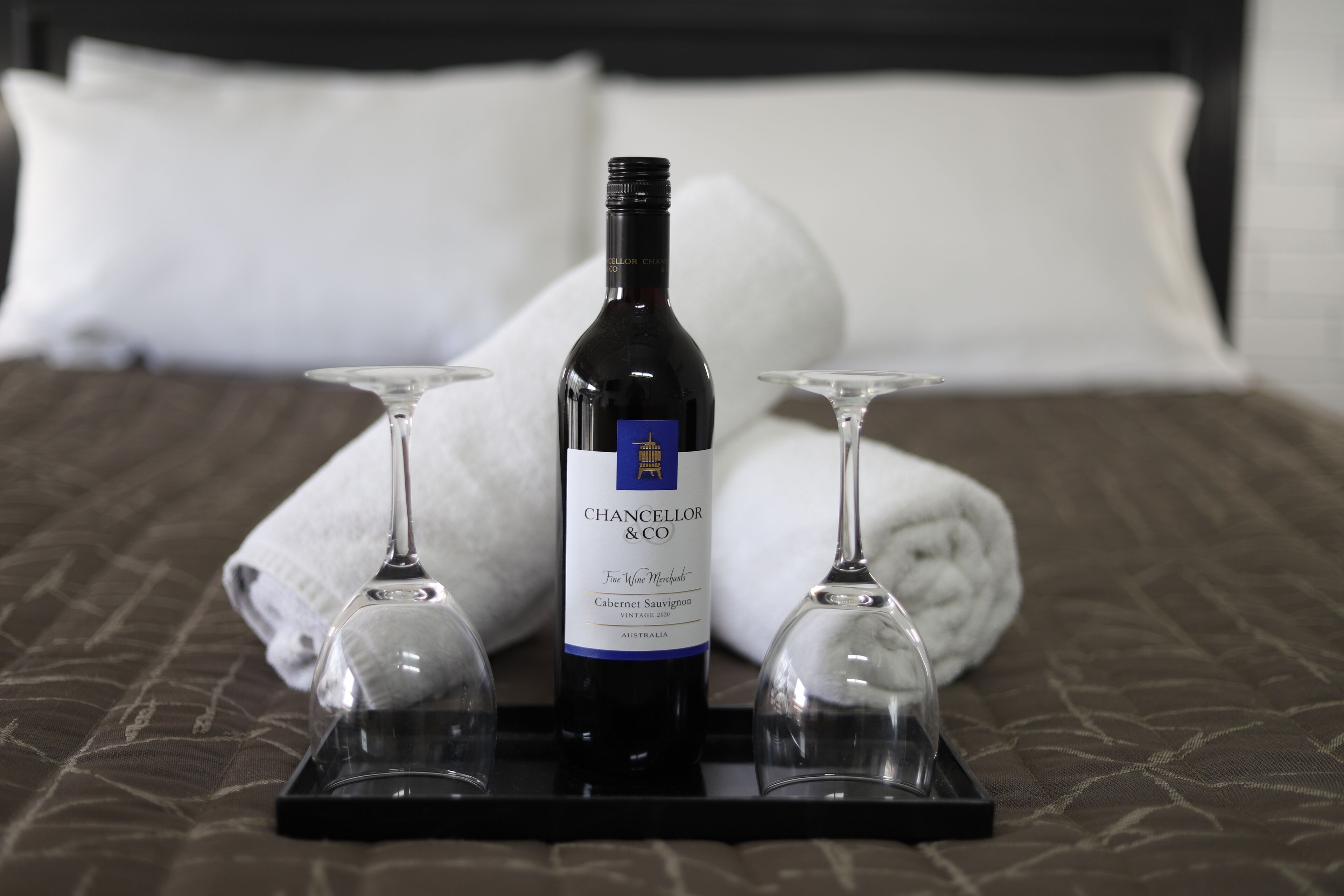 A bottle of Chancellor & Co. Cabernet Sauvignon on a tray with two wine glasses, placed on a bed with a brown bedspread and white pillows.