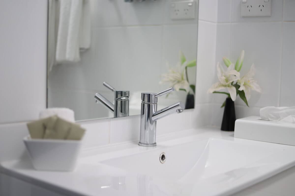A modern bathroom with a chrome faucet, a white sink, and a mirror reflecting a towel and a vase of white flowers.
