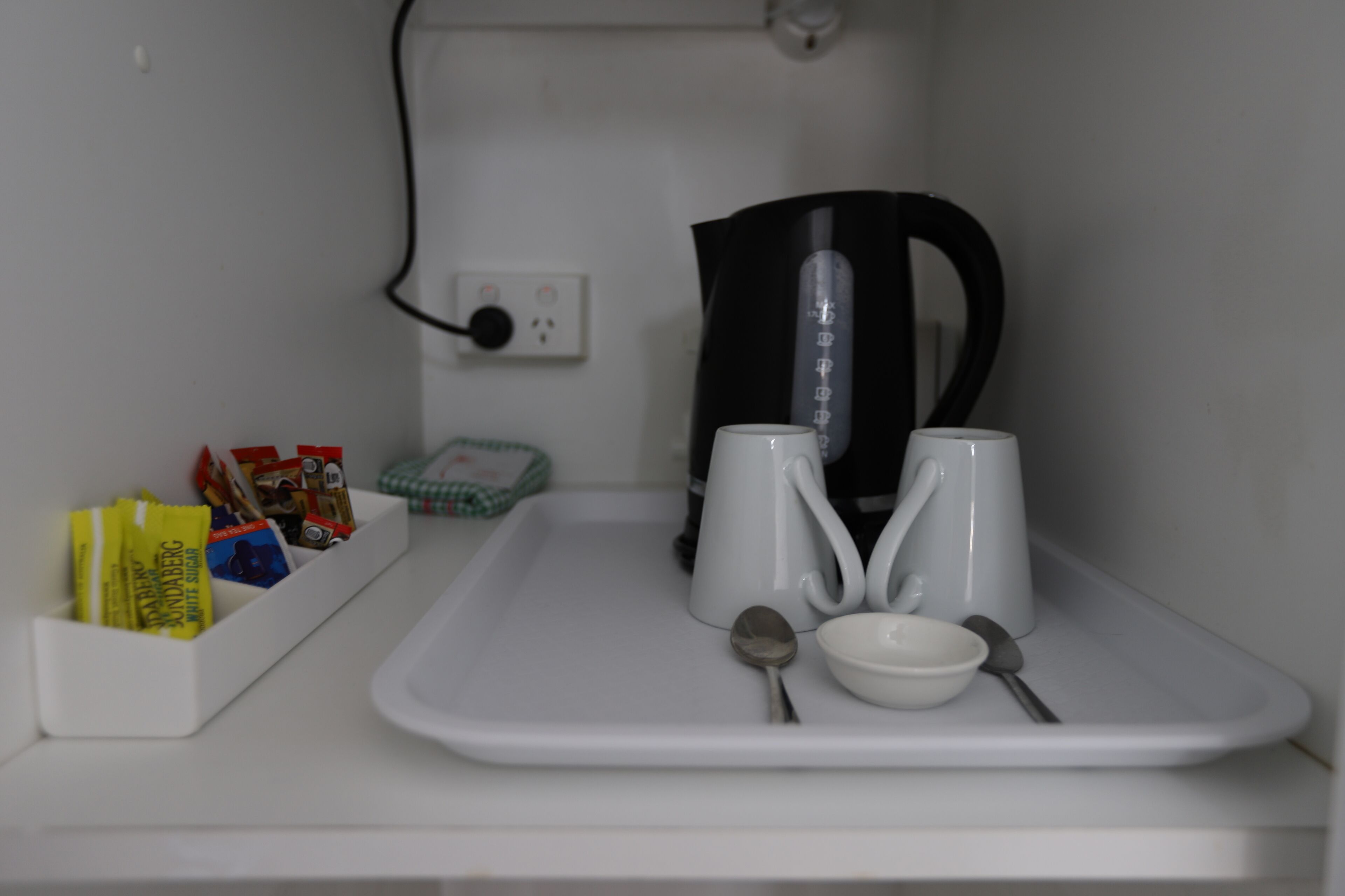 Una bandeja blanca con una tetera, dos tazas blancas, una cuchara y un bol. También hay sobres de azúcar y té a la izquierda.