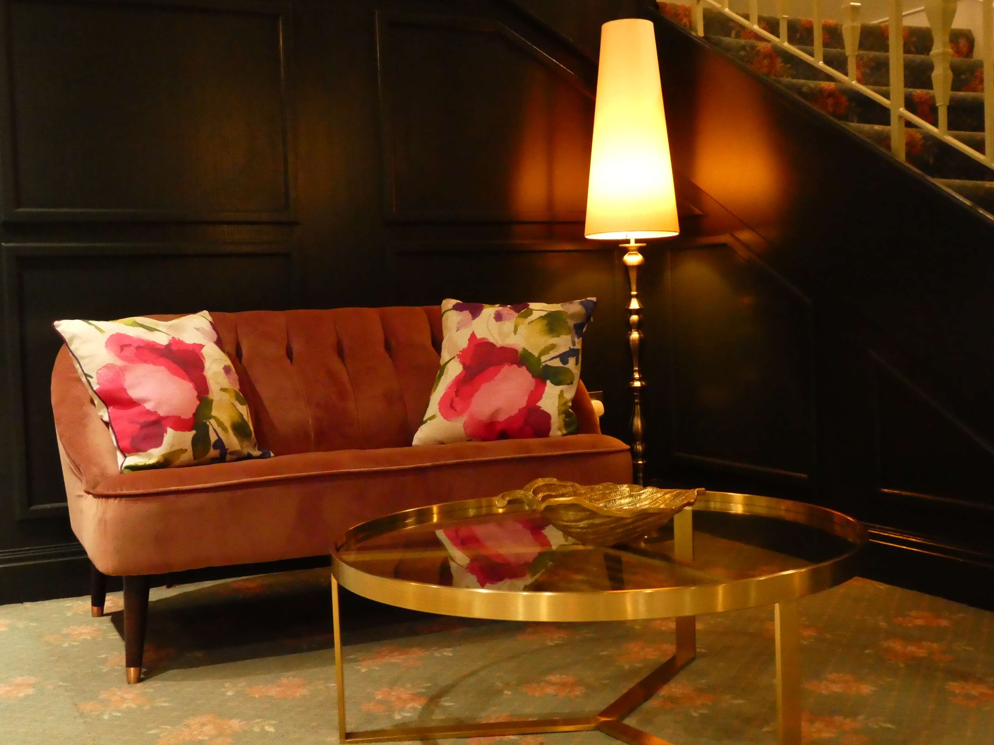 A brown sofa with floral cushions, a glass coffee table, and a lamp in a room with dark walls and a staircase in the background.