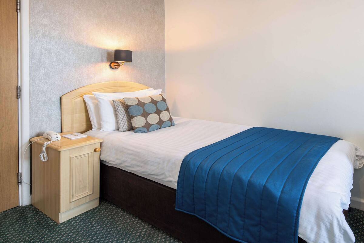 A hotel room with a bed, bedside table, lamp, and a dresser.