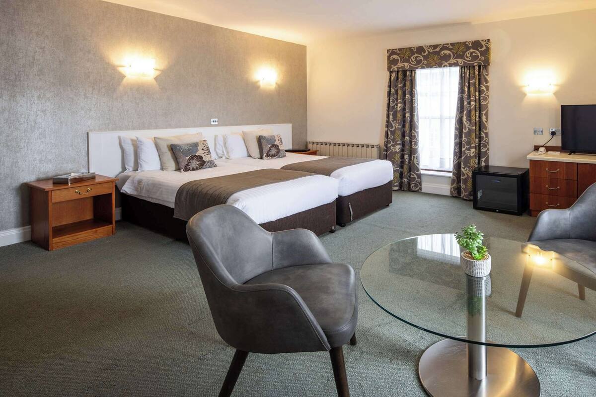 A hotel room with a bed, two armchairs, a glass coffee table, a TV, and a desk with a lamp.