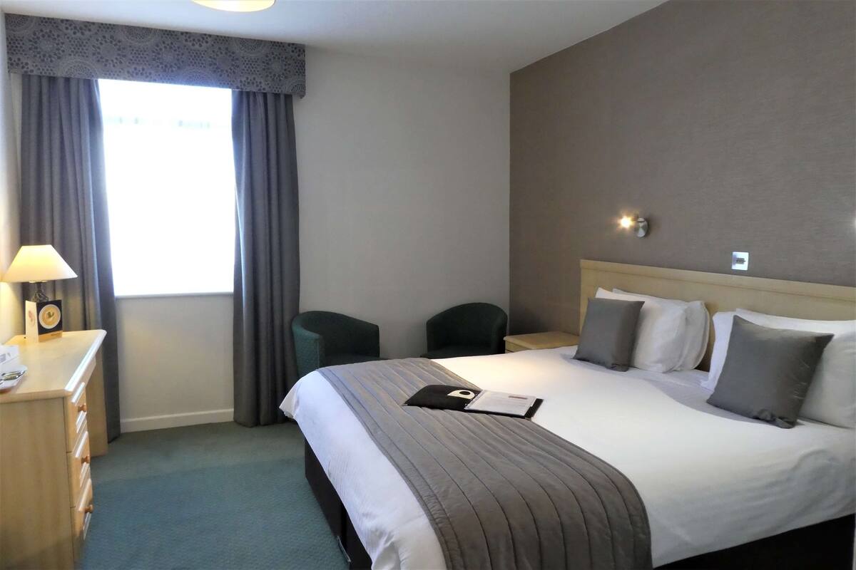 A hotel room with a large bed, a desk with a lamp, two chairs, and a window with curtains.