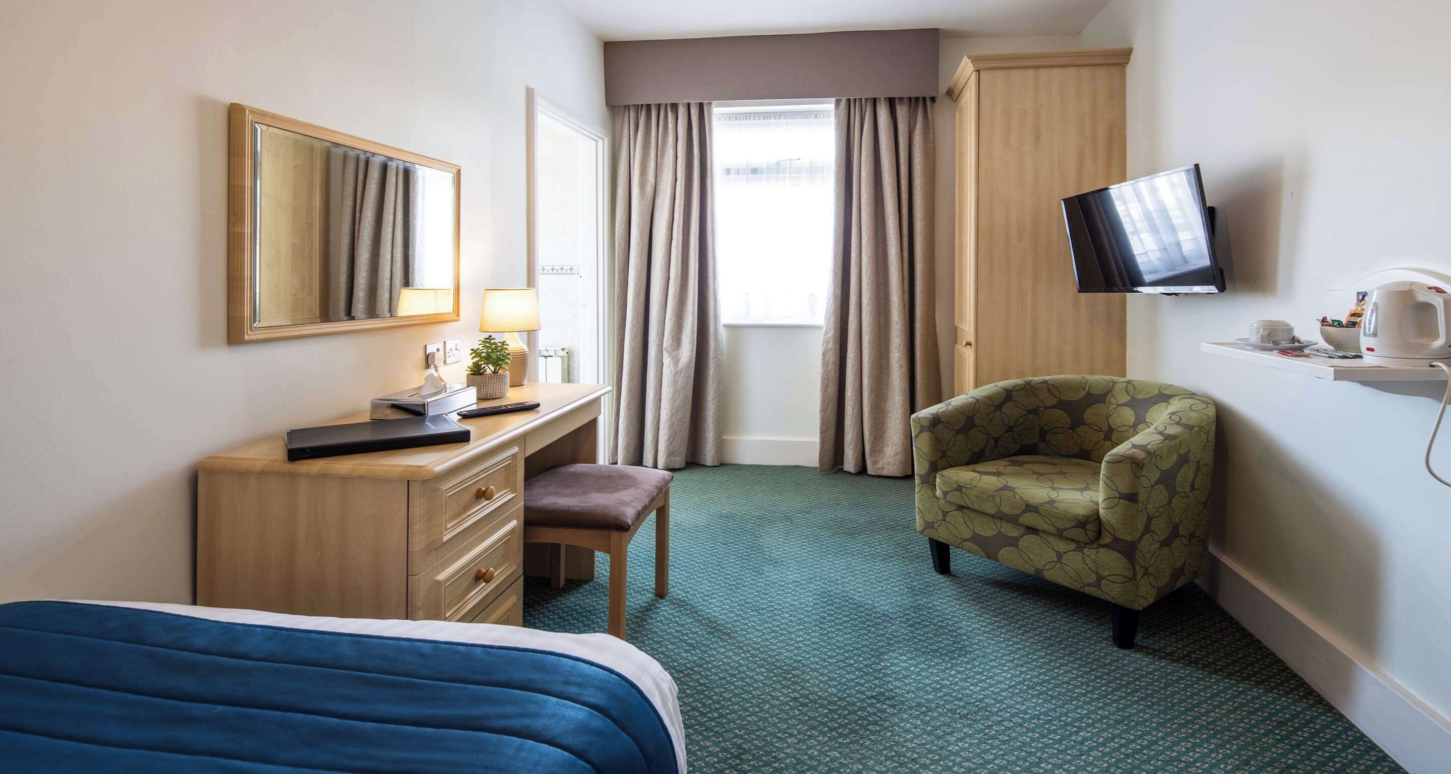 Una habitación de hotel con cama, escritorio con lámpara, silla, televisión y ventana con cortinas.