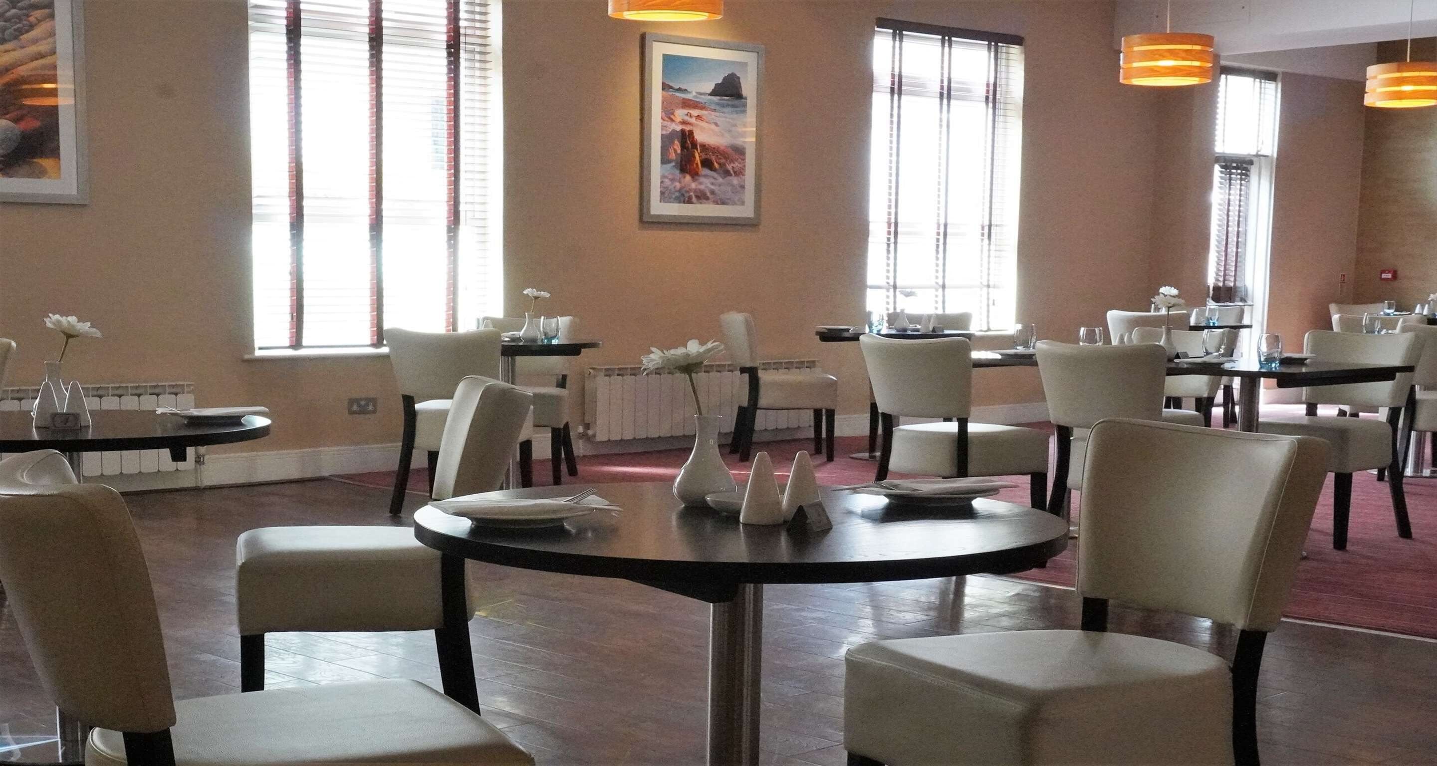 A restaurant with round tables and white chairs, large windows with curtains, and a framed picture on the wall.