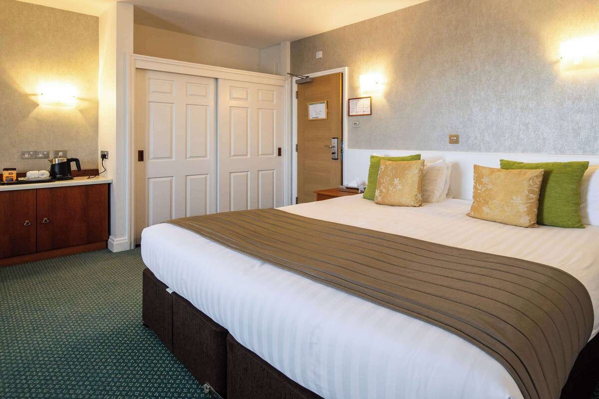 A hotel room with a large bed, a bedside table, a nightstand, a wardrobe, and a wall-mounted light fixture.