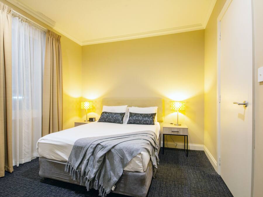 A hotel room with a bed, two bedside lamps, a nightstand, a door, and a thermostat on the wall.