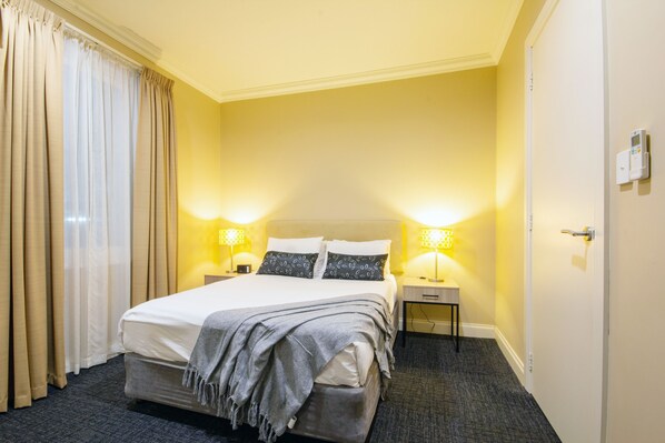 A hotel room with a bed, two bedside lamps, a nightstand, a door, and a thermostat on the wall.