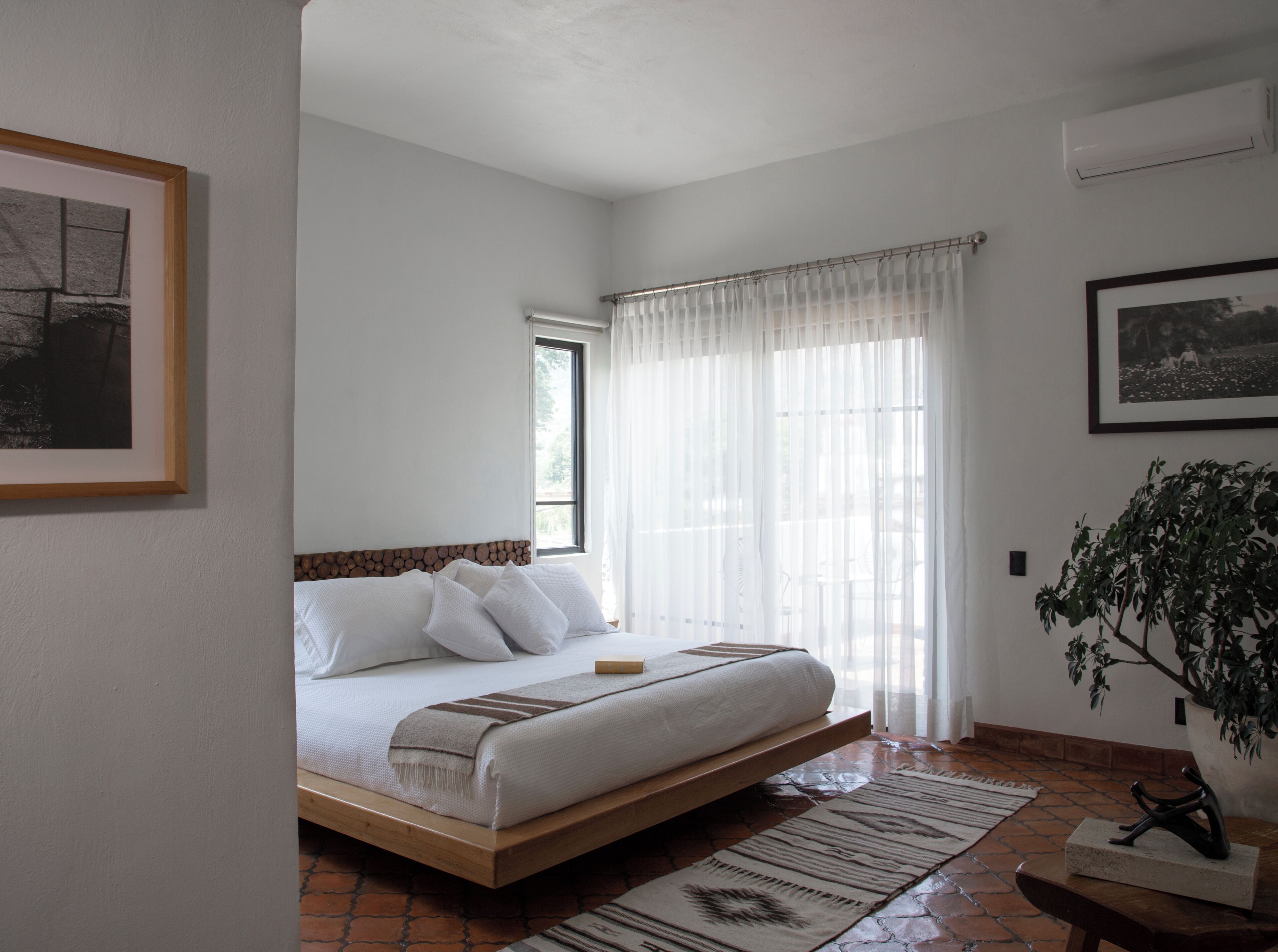 A bedroom with a bed, a window with curtains, a potted plant, and a framed picture on the wall.
