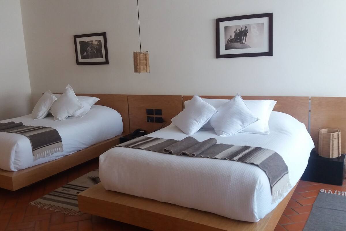 Une chambre d’hôtel avec deux lits simples, une tête de lit en bois, une petite table de chevet et une étagère, ainsi qu’un tableau accroché au mur.