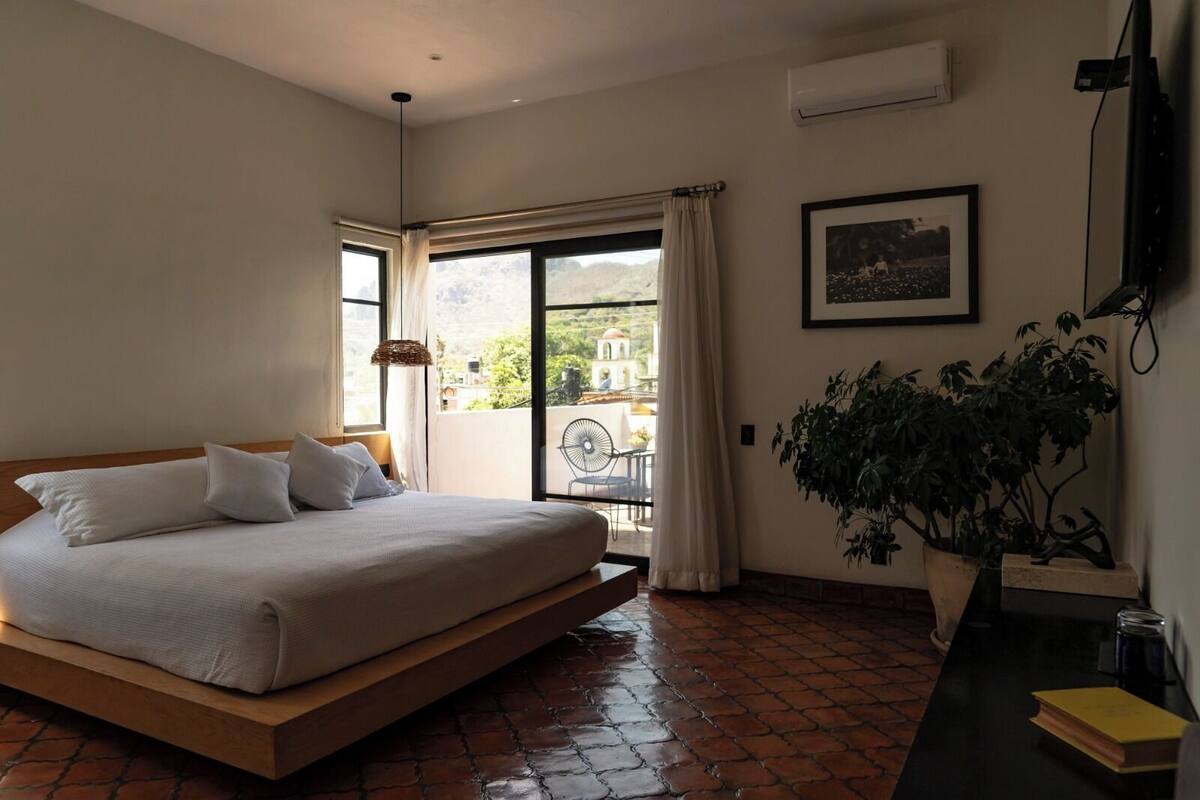 Une chambre avec un grand lit, un balcon avec une chaise, une plante en pot et un tableau encadré au mur.