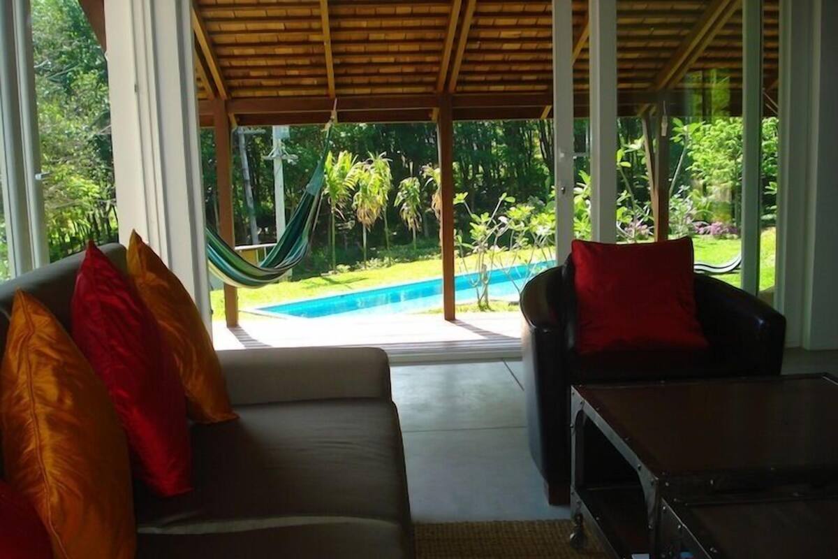 Una stanza con divano e poltrona, un'amaca e vista su una piscina e aree verdi.