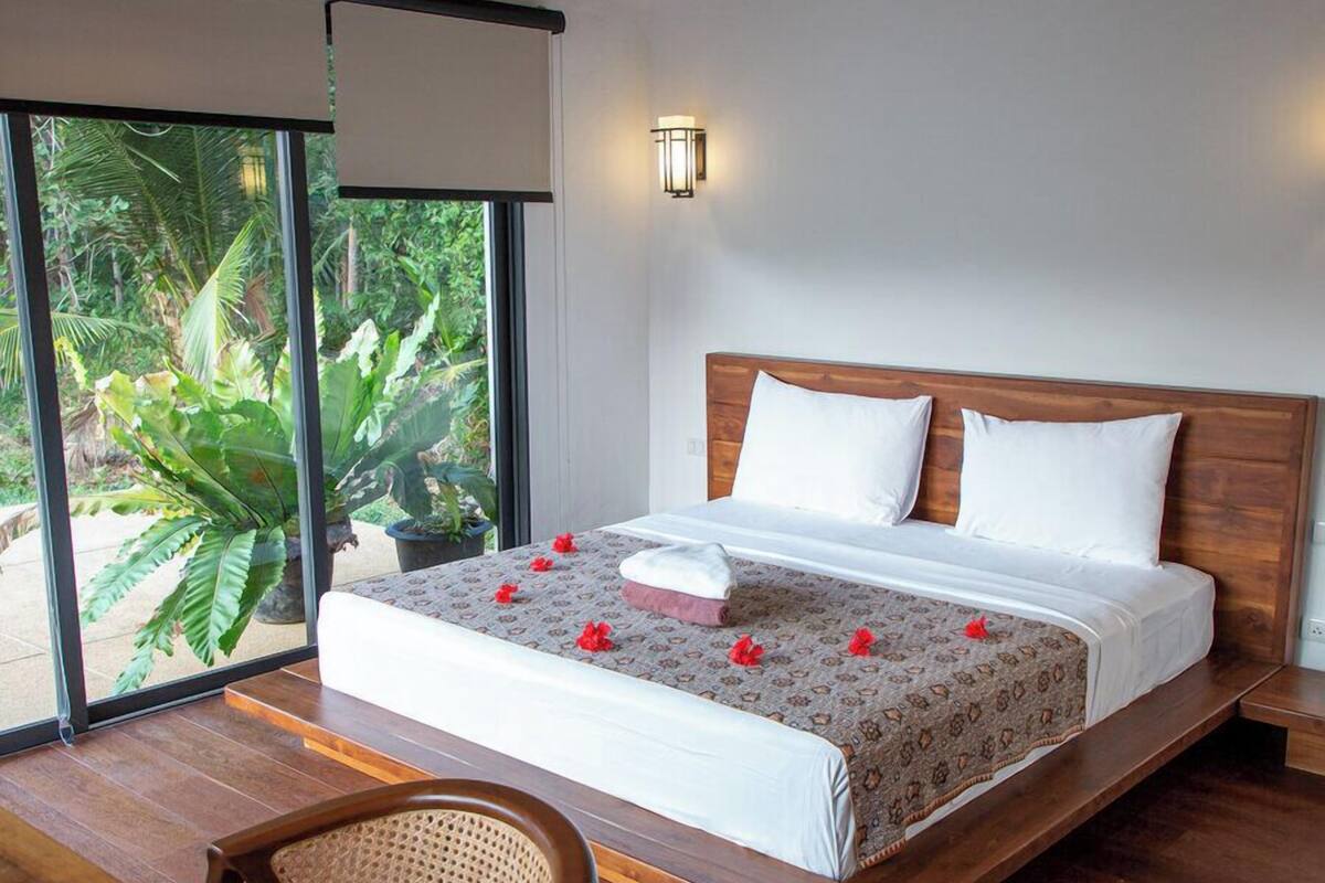 Camera da letto moderna con un letto in legno, biancheria da letto bianca e vista su una vegetazione lussureggiante attraverso ampie finestre.