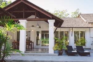 Un espacio al aire libre cubierto con sillones, plantas en macetas, una terraza de madera y un edificio con ventanas amplias y techo de tejas.
