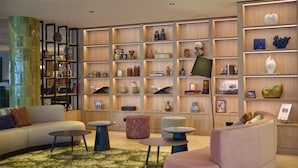 Ein modernes Wohnzimmer mit einer Couch, zwei runden Tischen und einem rosafarbenen Sessel. Im Hintergrund steht ein Holzregal, das mit dekorativen Gegenständen und Büchern gefüllt ist.