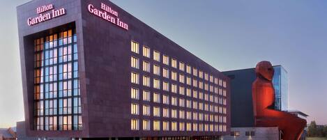 Un Hilton Garden Inn de diseño moderno, con un amplio frente de vidrio y una escultura roja distintiva a la derecha.