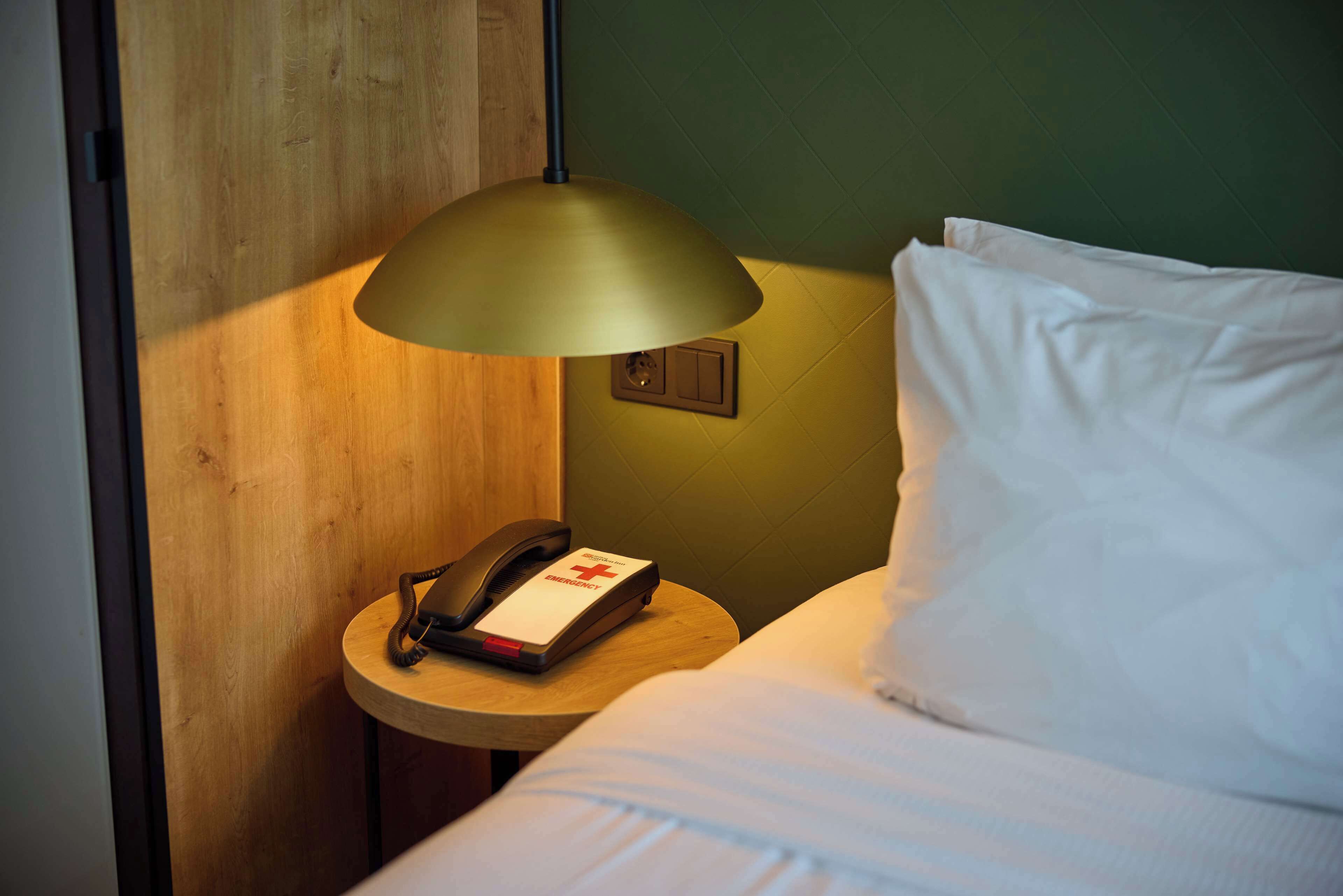 A hotel room with a bed, a bedside table with a lamp, a phone, and an emergency button.