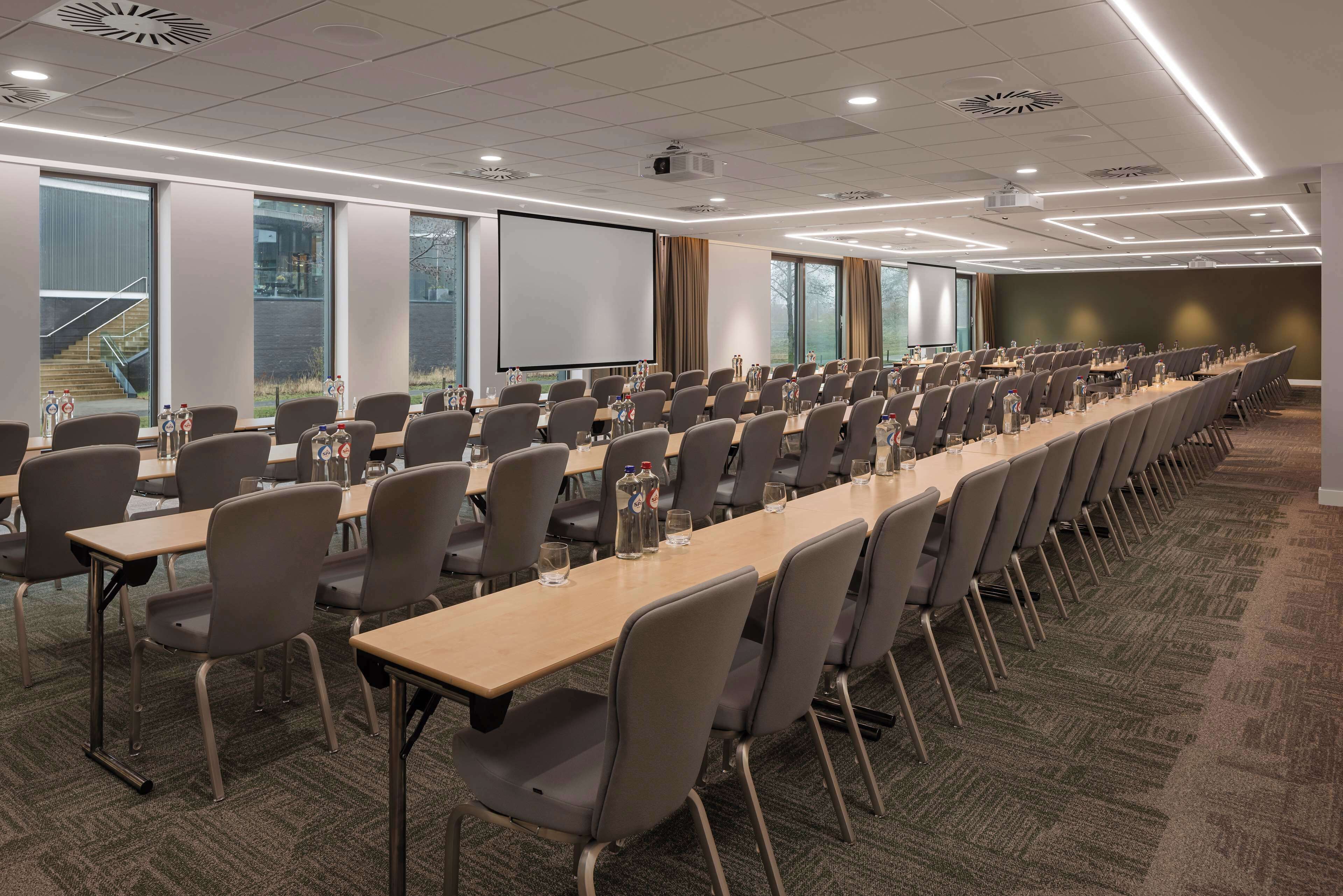 Uma sala de conferências com fileiras de mesas e cadeiras, janelas amplas e uma tela de projetor.