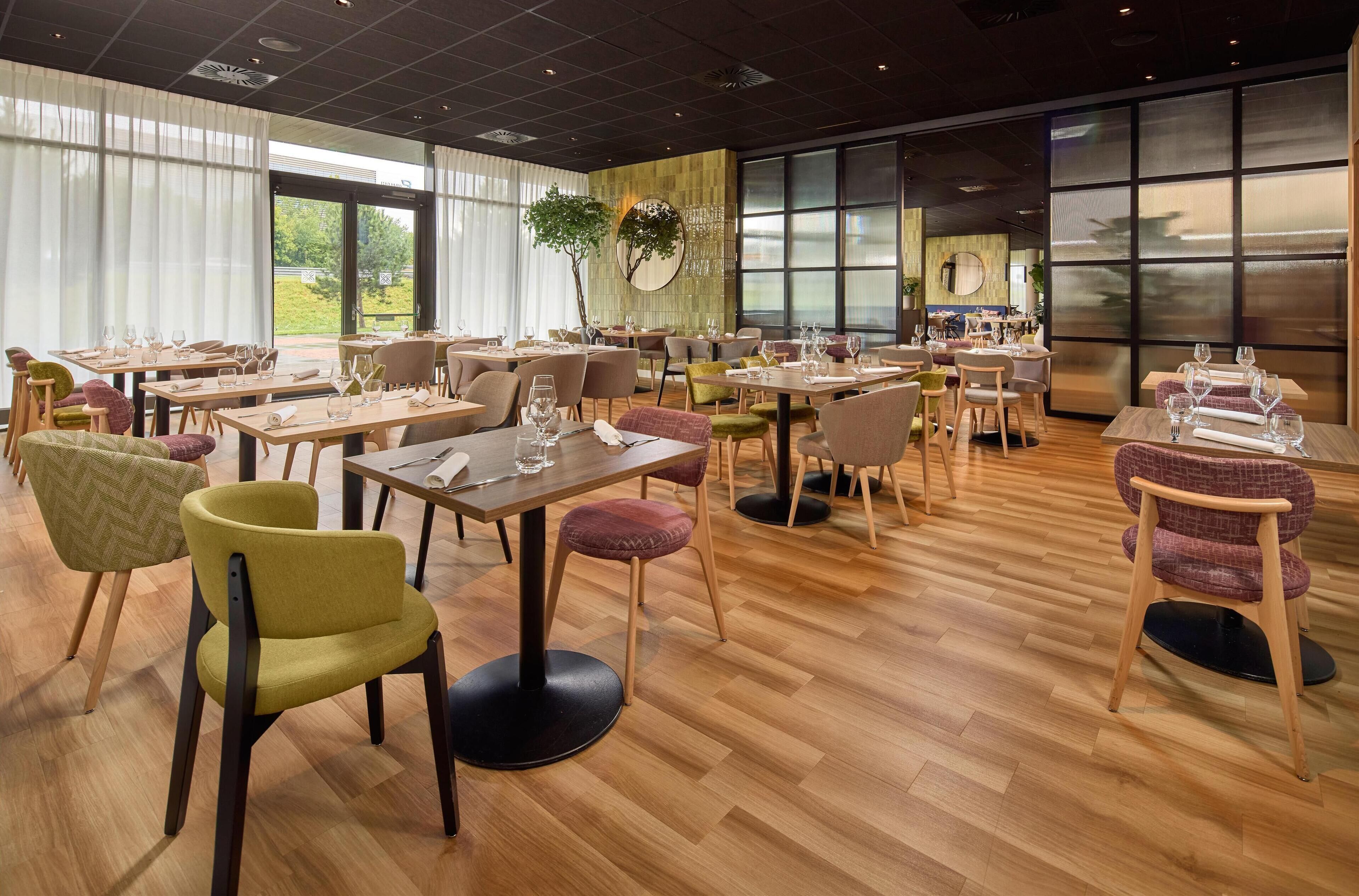 A restaurant with multiple tables and chairs, large windows, and a plant in the center.