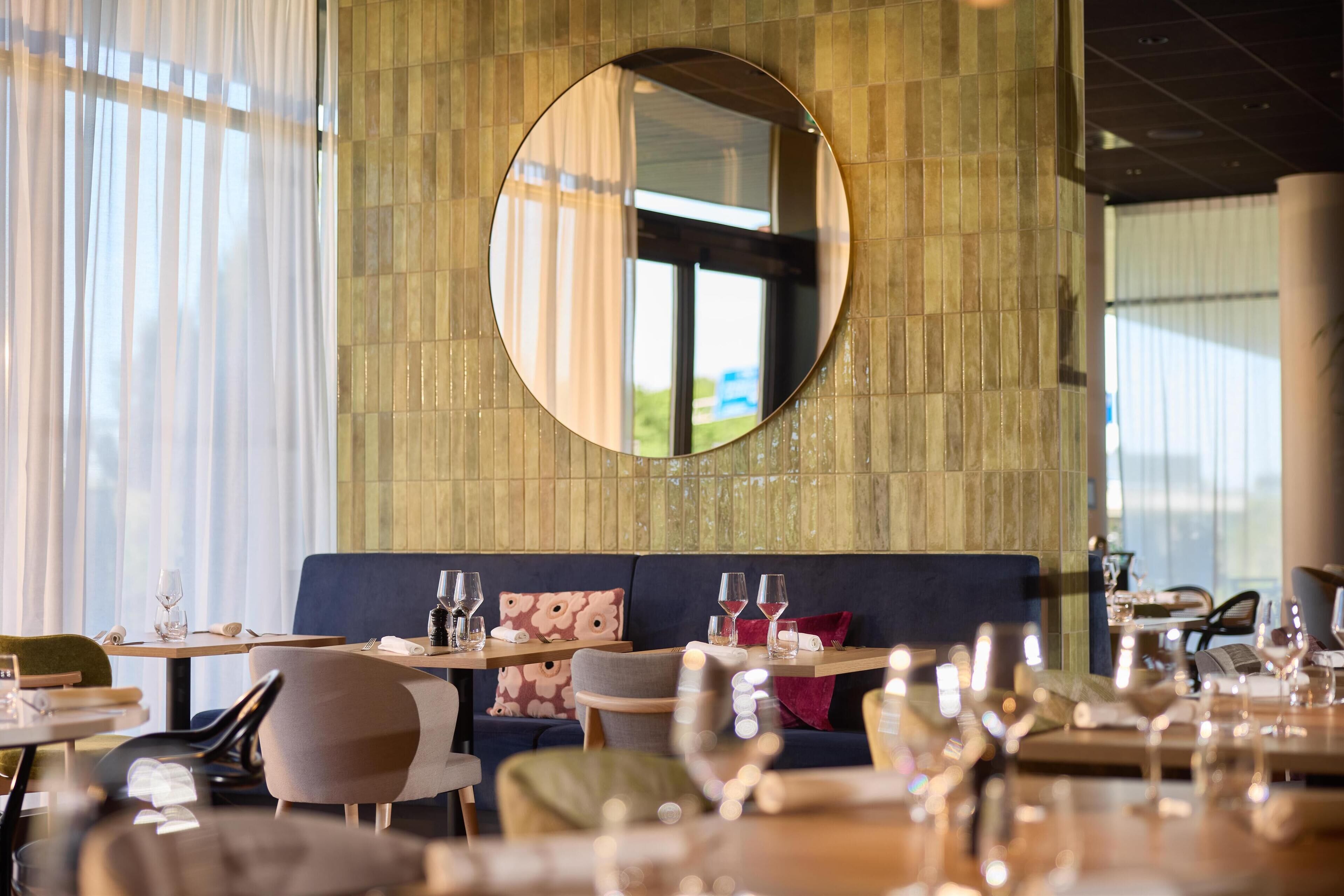 Interior de restaurante con mesas redondas, asientos acolchados y un espejo grande en la pared.