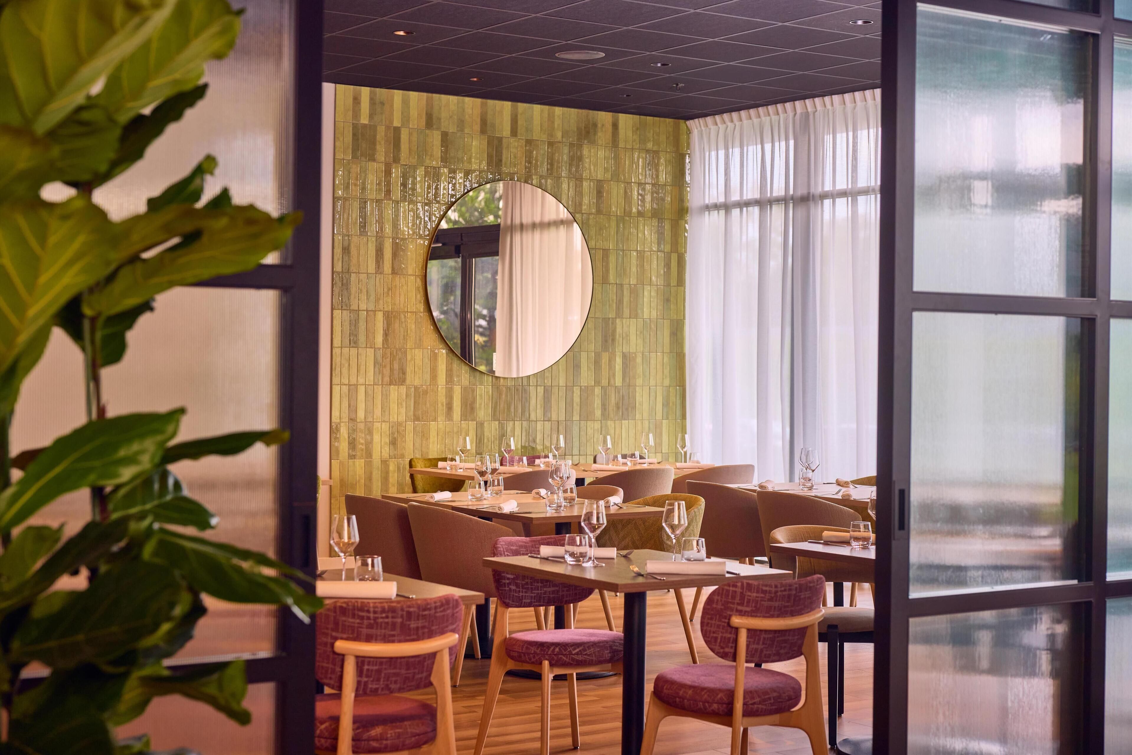 A restaurant with a modern interior, featuring a green wall with a mirror, wooden tables, and purple upholstered chairs.