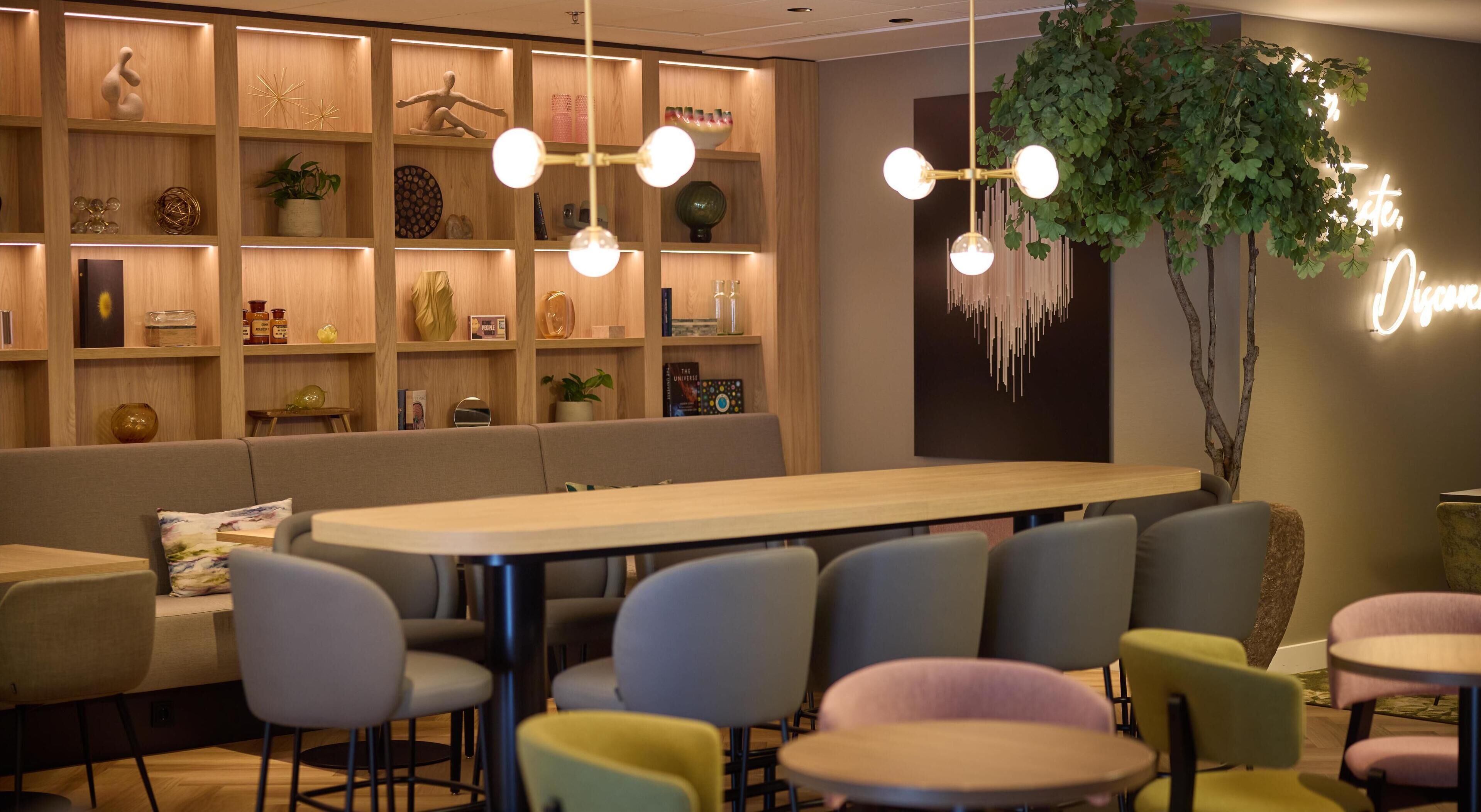 A modern dining area with a long wooden table, multiple chairs, and a bookshelf filled with various decorative items.