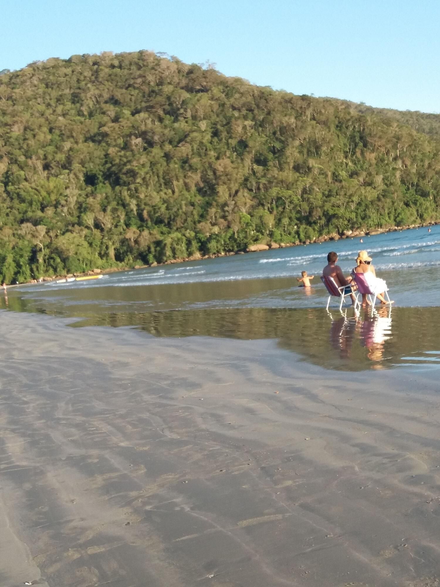 A beach with clear water and a forested hill in the background.