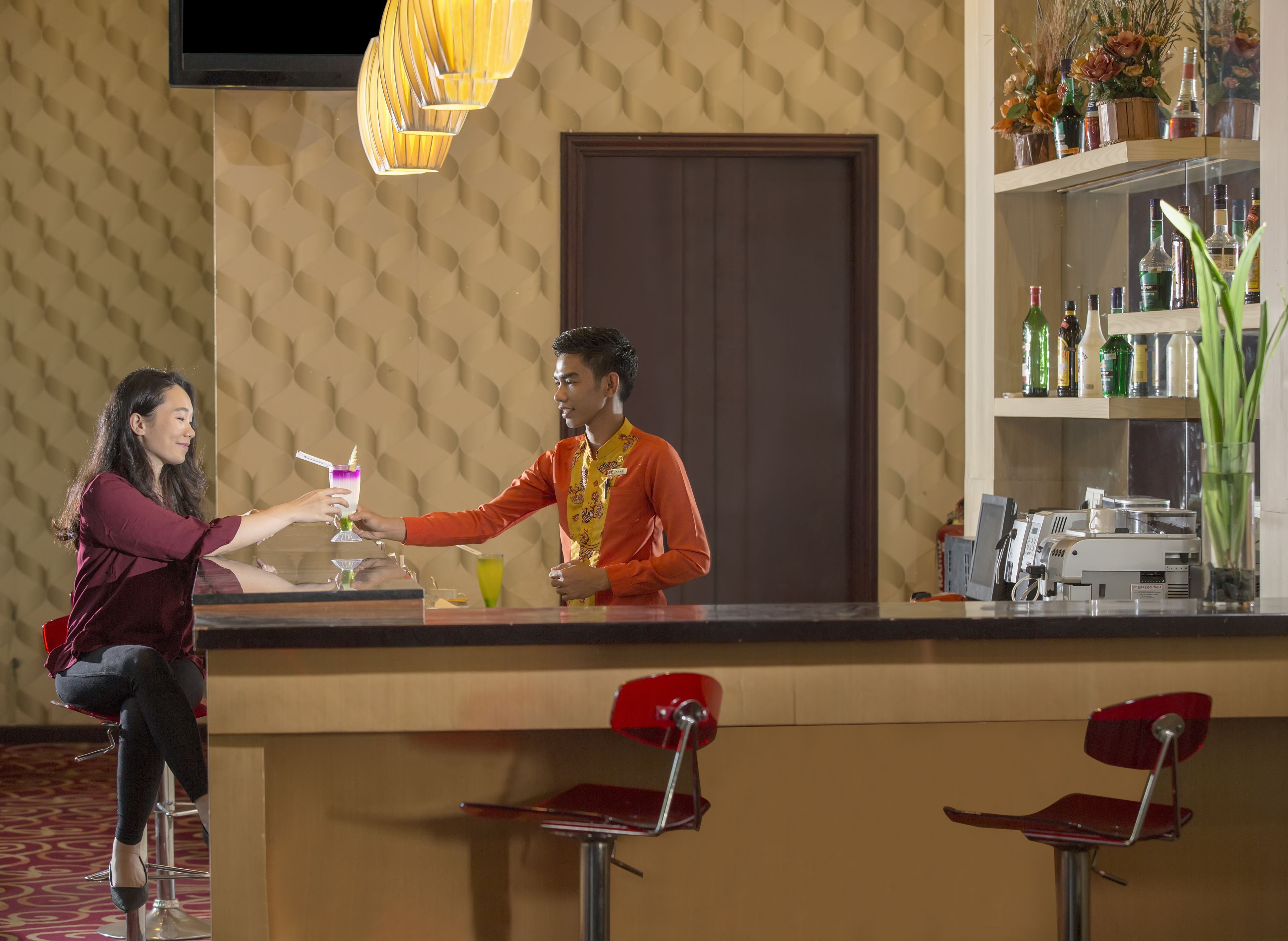 Angolo bar con un barista che serve un drink a un cliente. Sono presenti sgabelli da bar, un bancone e scaffali pieni di bottiglie.