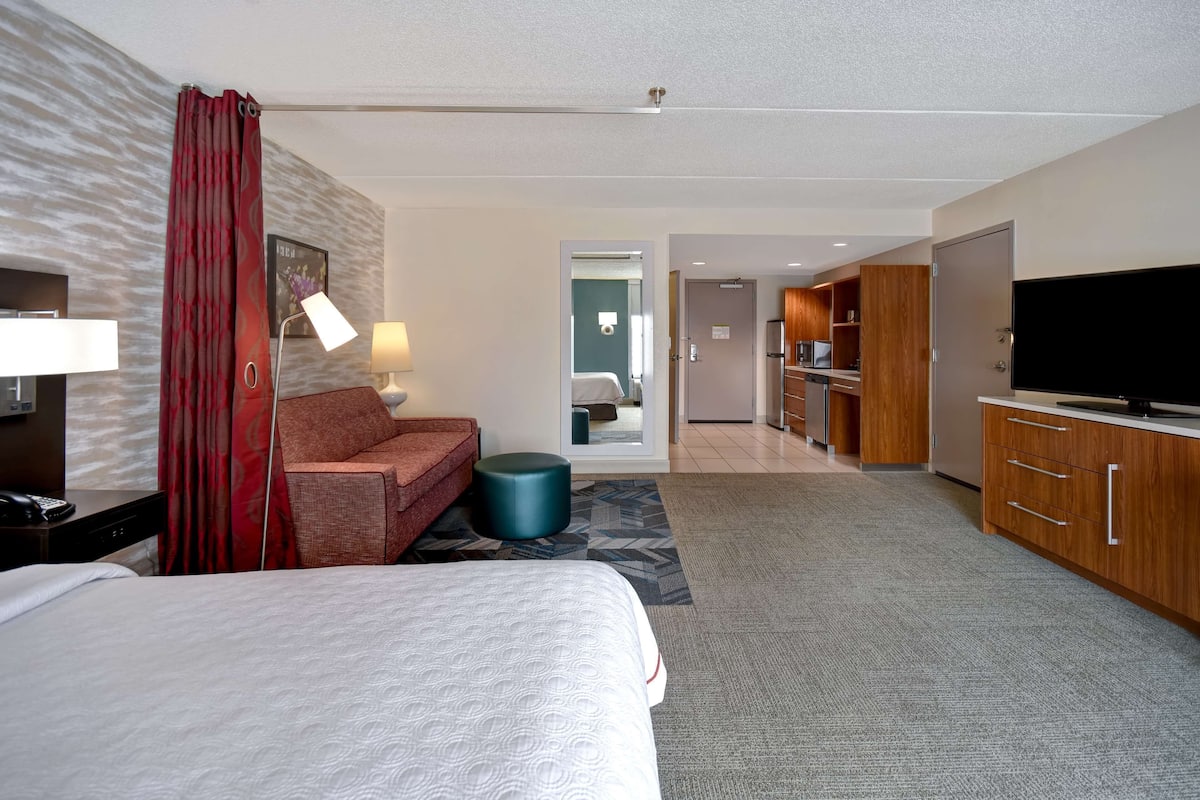 Habitación de hotel con cama, sofá, televisión, escritorio y baño visible a través de una puerta abierta.