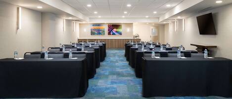Un salón de conferencias con filas de mesas, botellas de agua y un cuadro grande en la pared.