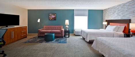 A hotel room with a flat-screen TV, a sofa, two beds, a desk, and a small table with lamps.