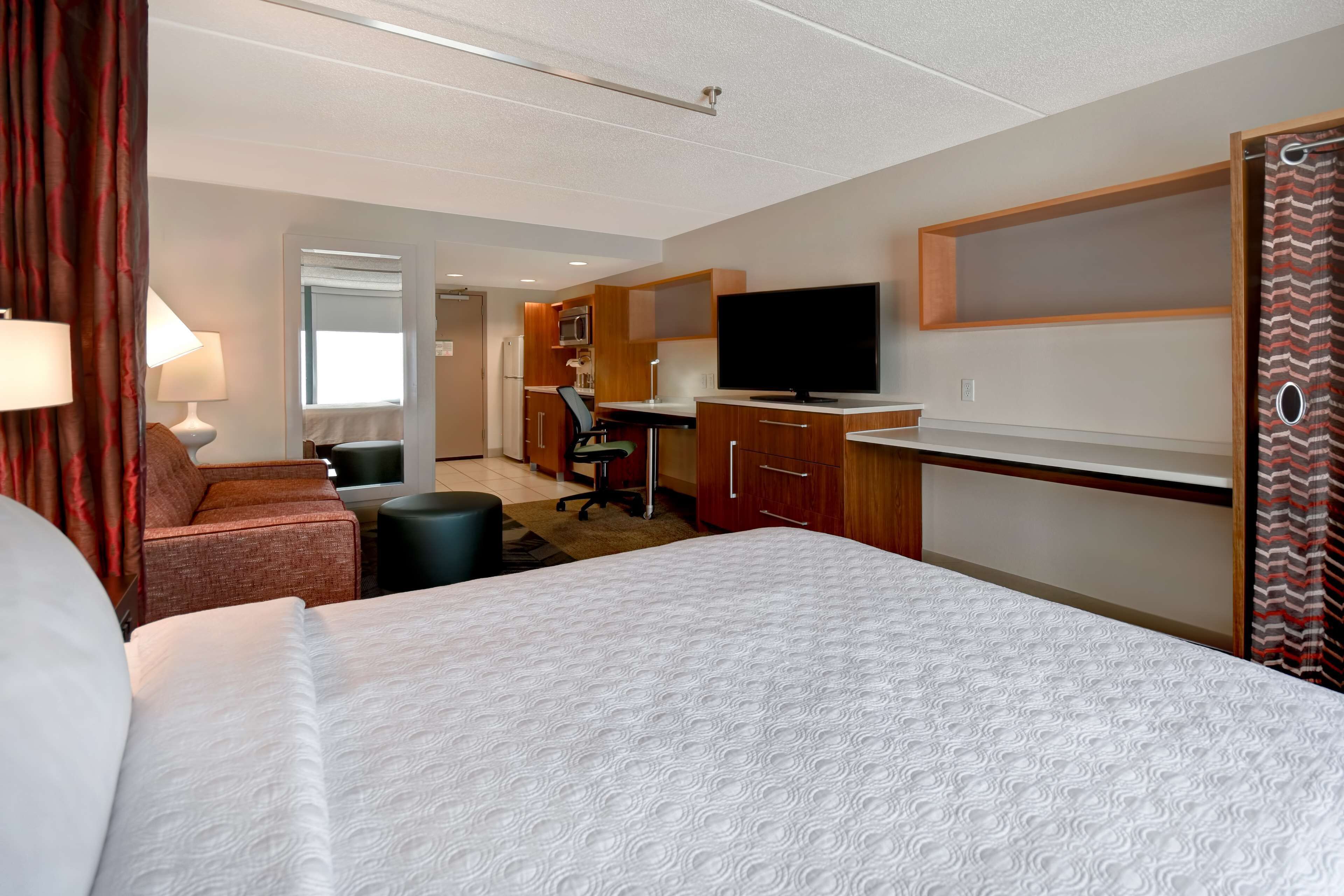 A hotel room with a large bed, a desk with a flat-screen TV, a kitchenette, and a seating area with a chair and a small table.