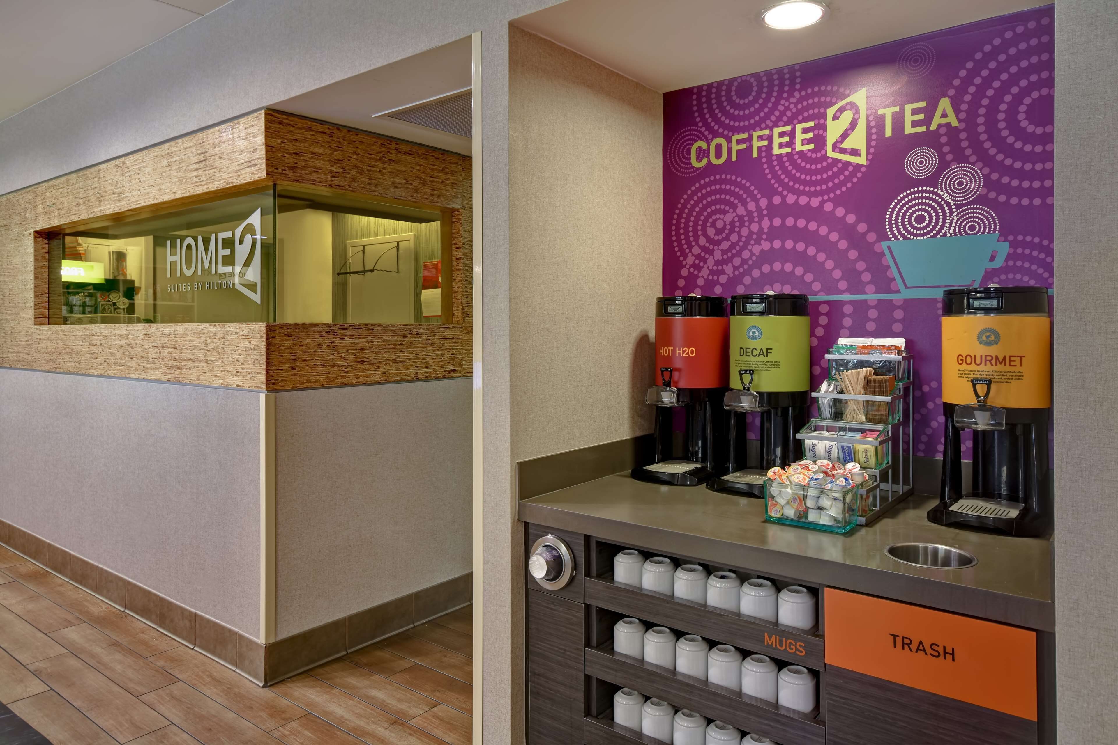 A hotel lobby with a coffee station featuring a 'Coffee 2 Tea' sign, a 'Home 2 Suites by Hilton' sign, and a trash bin.