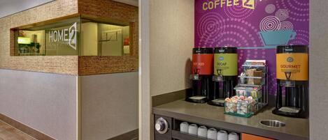 A hotel lobby with a coffee station featuring a 'Coffee 2 Tea' sign, a 'Home 2 Suites by Hilton' sign, and a trash bin.